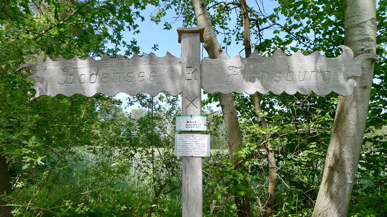 Hölzerner Wegweiser am Europäischen Fernwanderweg E1. Ein mit „Bodensee“ beschriftetes Schild zeigt nach links, ein mit „Flensburg“ beschriftetes Schild nach rechts.