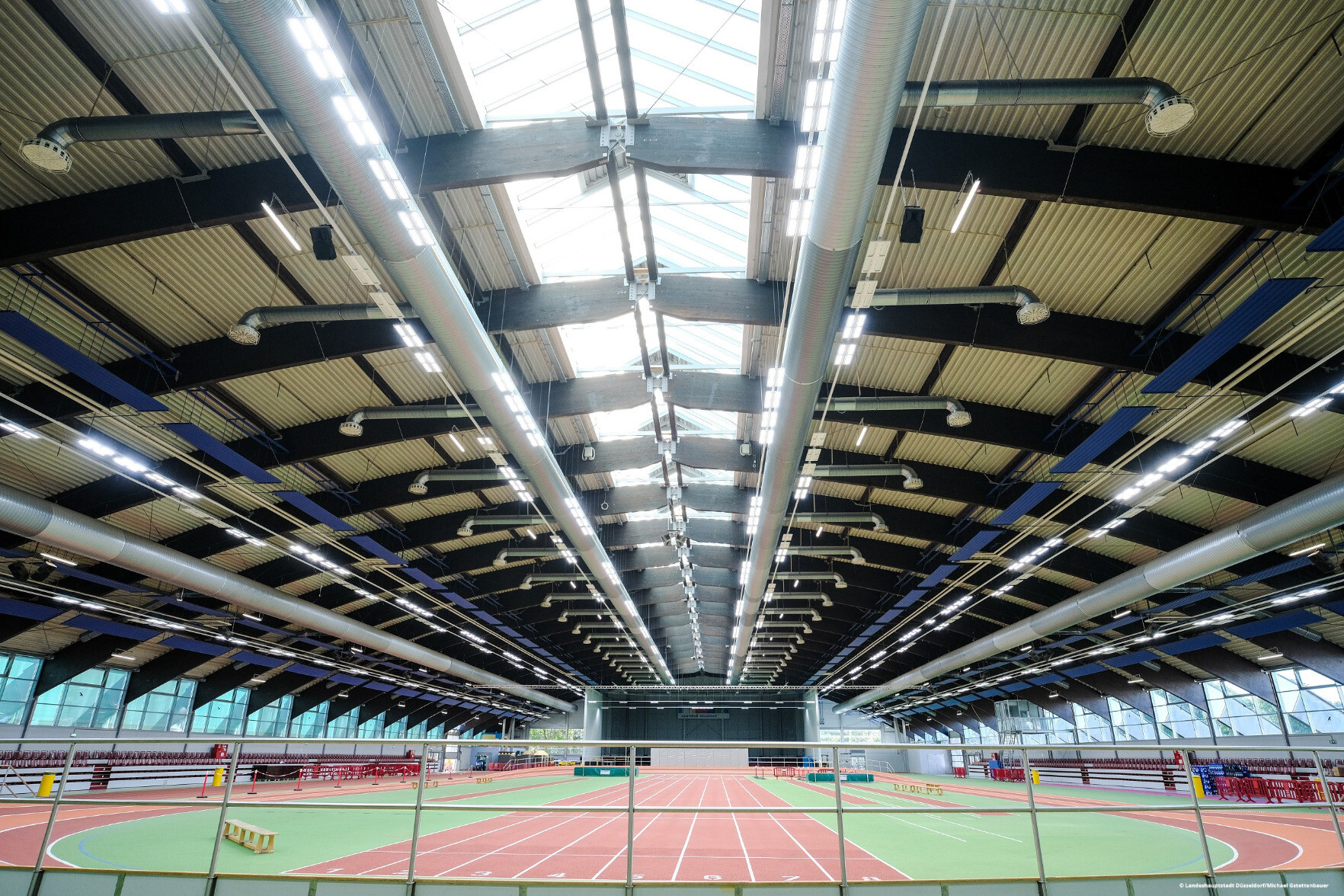 Blick in die Leichtathletikhalle im Arena-Sportpark in Düsseldorf. Wir schauen in Richtung Decke und sehen die neuen LED-Leuchten. Darunter sind ein Sportfeld und Tribünen zu erkennen. 