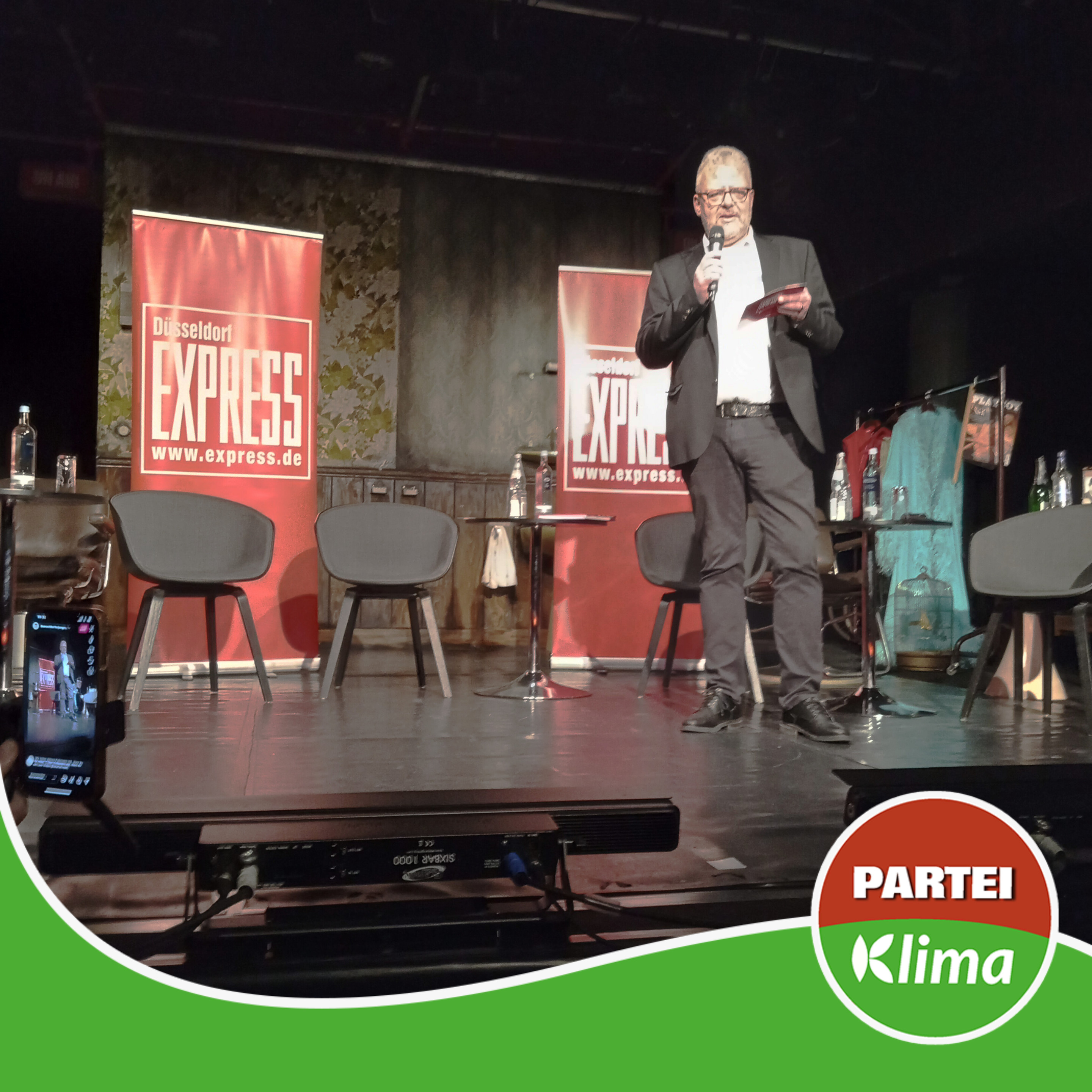 Zu sehen ist Herr Michael Kerst, der Moderator des EXPRESSO Talks der Düsseldorfer EXPRESS Redaktion auf der Bühne des Theaters an der Kö. Hinter ihm befinden sich eine Stuhlreihe aus 5 Stühlen und die Banner des EXPRESS.