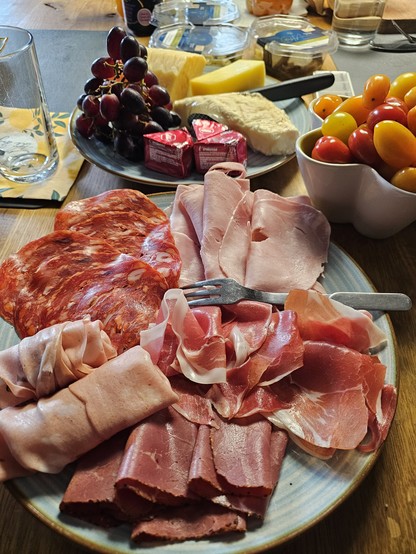 Italienischen Wurst- und Schinkensorten auf einem Teller angerichtet, dahinter ein Teller mit Käse und Weintrauben, daneben ein Schälchen mit Tomaten 