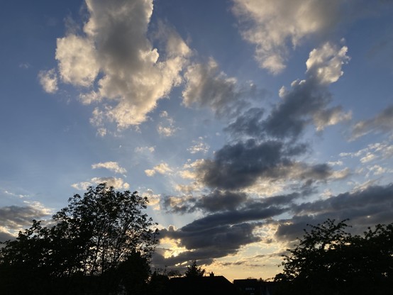 Am unteren Bildrand die Silhouetten von Bäumen (mittig links und rechts je eines großen Baumes), Sträuchern und Dächern. Mittig unten zeigt sich das letzte Licht der untergehenden Sonne, die selbst nicht zu sehen ist. Die Sonne sendet helles Licht über den Himmel. Der Himmel ist teilweise mit flauschig grauen und weißen Wolken bewölkt.