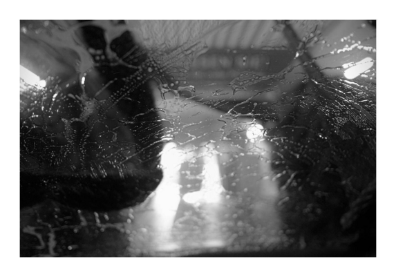 Wasser und Reinigungsmittel auf einer Scheibe, im Hintergrund unscharf Konturen einer Waschstraße, schwarz-weiß-Bild.