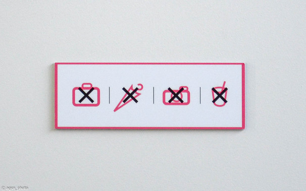 Eine kleine Verbotstafel an einer weißen Wand in einem Museum. 
Die vier roten Symbole für Koffer, Regenschirm, Kamera und Trinkbecher sind alle mit einem fetten schwarzen X durchgestrichen.
