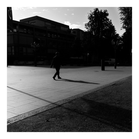 Ein Herr in einem dunklen Anzug läuft morgens bei tief stehender Sonne auf einen Schatten eines großen Gebäudes zu. Der Oberkörper ist vor dem dunklen Hintergrund des Gebäudes kaum zu sehen. Quadratisches schwarz-weiß Foto.