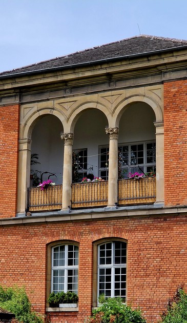 Balkon eines Backsteinhauses. Der Balkon hat vier Säulen, dazwischen sind Metallgestelle mit einer Art Holzverkleidung, darüber Blühpflanzen. Im Hintergrund sind Sprossenfenster zu erkennen.