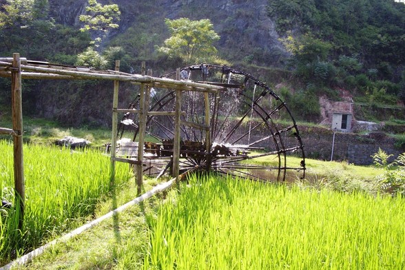 A large waterwheel in a lush green field with trees and hills in the background.