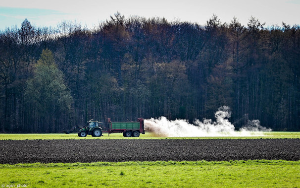 Ein Traktor fährt auf einer Wiese und streut von seinem Anhänger Dünger aus, der weißen Rauch bildet.