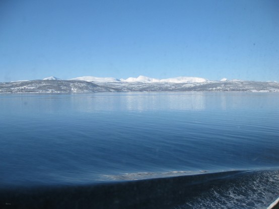 A serene landscape featuring a calm blue lake with snowy hills and mountains in the background under a clear blue sky.