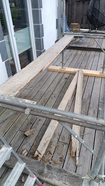 Vor einer Terrassentür auf einer Dielenterrasse, Stahlgerüstrahmen, zuunterst  halbhohe Rahmen. Die etwa 45 Zentimeter breite Bohlen ist zur Hälfte gebrochen, die Bruchstücke liegen am Boden