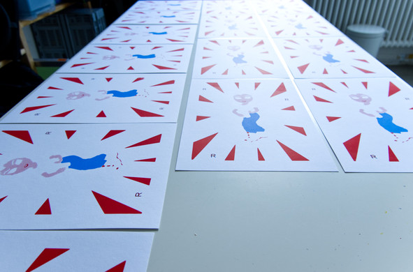 Viele identische Drucke liegen auf einem Tisch und zeigen eine unfertige Figur aus hellblau und rot sowie rote Zacken, die die Figur umgeben wie ein Kranz. 