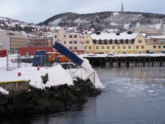 Snow-covered dock with a blue truck unloading snow into the water, in front of yellow buildings and a mountainous background.