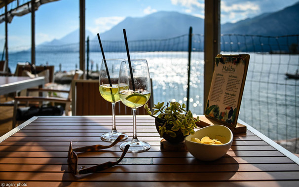 Auf einem kleinen Tisch in einem Strandcafé stehen zwei hohe Weingläser mit einem Zitronengetränk. Dahinter erkennt man in der Unschärfe einen See und ein hohes Gebirge.