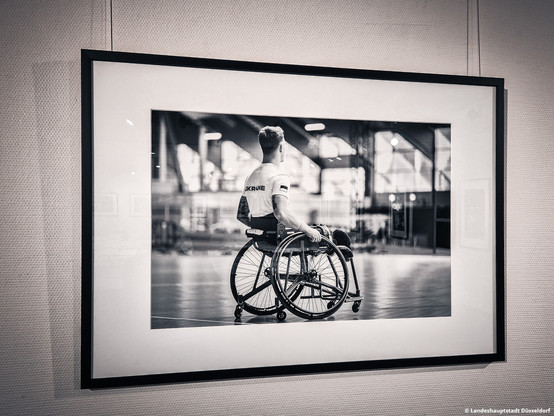 Ein Foto der Austellung zu den Invictus Games in Düsseldorf. Das Schwarz-Weiß-Bild zeigt einen Athleten im Rollstuhl. Es hängt in einem schwarzen Rahmen an einer weißen Wand. 