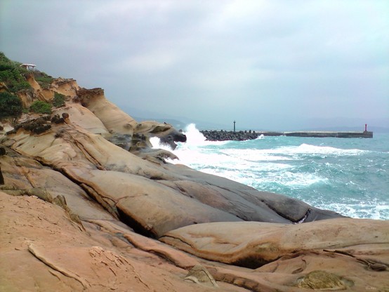 Rocky coastline with unique rock formations, waves crashing against the shore, and a jetty extending into the sea under an overcast sky.