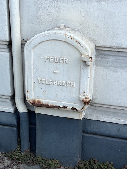 Grauweisser, angerosteter Metallkasten an einem Haus in ca 40 cm Höhe mit der hervorstehenden Beschriftung 