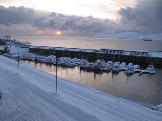 Snow-covered harbor with docked boats, snowy ground, lampposts, and mountains in the background during sunset.