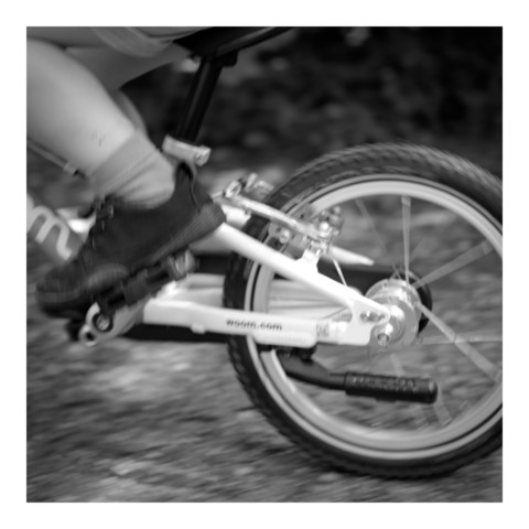 Schwarz-weiß Foto eines Kinderrads mit teilweise sichtbarem Fuß und Bein des Kindes. Ansonsten sind nur das Hinterrad (angeschnitten) und ein Teil des Rahmens sichtbar. Hintergrund und Teile des Rads sind aufgrund des Bewegung unscharf. 