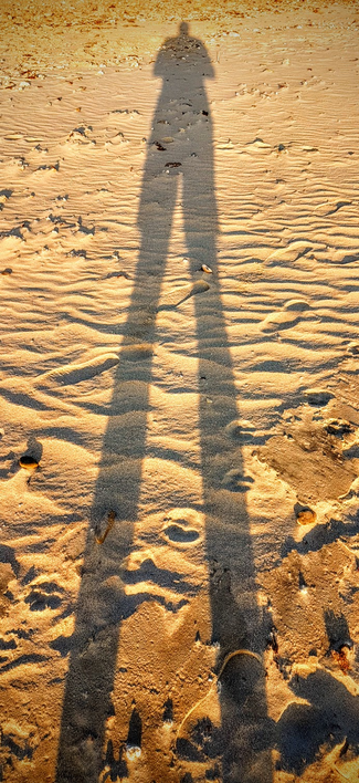 Ein menschlicher Schatten fällt im Sonnenuntergang auf einen Sandstrand. Der Körper wirkt daher extrem gestreckt mit sehr langen Beinen... 