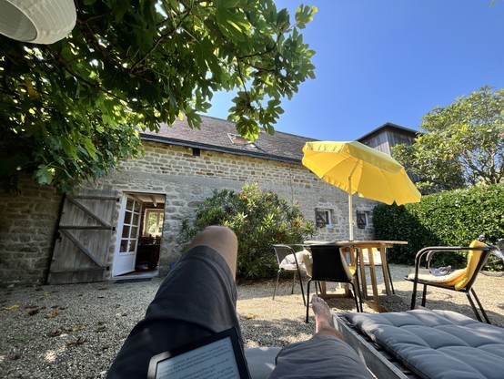 Person, die mit einem E-Reader in einem Garten liegt. Backsteingebäude mit offener Tür im Hintergrund. Gartenmöbel mit gelbem Regenschirm. Üppiges Grün und blauer Himmel im oberen Bildbereich.