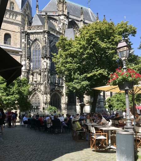 Menschen sitzen im Freien an den Tischen eines Cafés am Münsterplatz in Aachen. Die Tische und Stühle stehen im Schatten. Im Hintergrund ist ein Teil des Aachener Doms zu sehen, der von der Sonne angestrahlt wird. Rechts stehen ein großer Baum und eine Laterne, an der eine Blumenampel mit roten Blüten hängt. Der Himmel ist strahlend blau und wolkenlos.