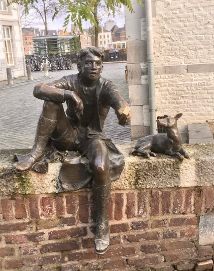 Bronzestatue eines Jungen, der auf einer Steinmauer sitzt und ein rechteckiges Stück Seife hält. Der Junge lässt ein Bein baumeln, das andere ruht angewinkelt auf der Mauer.  Neben ihm liegt ein kleiner Bronzehund, der zu ihm aufschaut. Im Hintergrund sieht man eine Straße mit Kopfsteinpflaster, Gebäude rechts und links und Fahrräder.