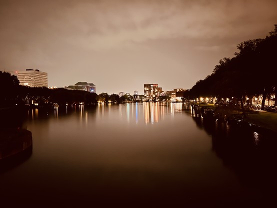 Amsterdam bei Nacht. Auf dem Weg zum Hotel über eine Brücke gelaufen. Der weite Blick über das Wasser. Rechts und links Bäume. Ganz weit hinten eine Skyline aus Häusern, die sich hell erleuchtet im Wasser spiegeln und schöne lange farbliche Striche im Wasser hinterlassen. Das Bild selbst ist in dunklen schwarz/braun-Tönen gehalten.