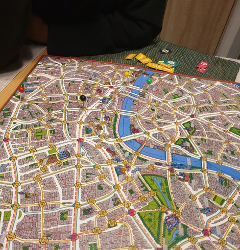 Spielfeld Scotland Yard. Plan von London, Taxi-, Bus- und U-Bahnrouten bunt eingezeichnet. Am Rand liegen einige Spiel-Chips für Taxifahrten, Bus und U-Bahntickets.