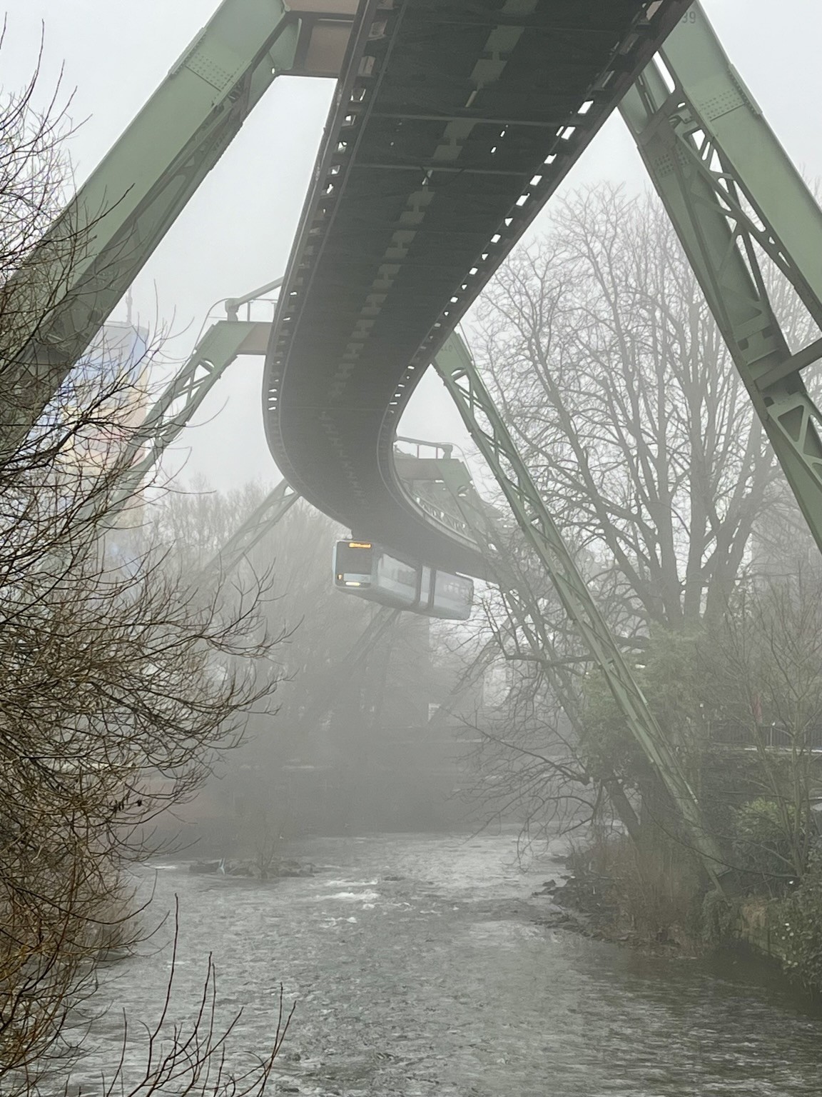 Schwebebahn über der Wupper, umgeben von Nebel und kahlen Bäumen. Die Szene hat eine gedämpfte, neblige Atmosphäre.